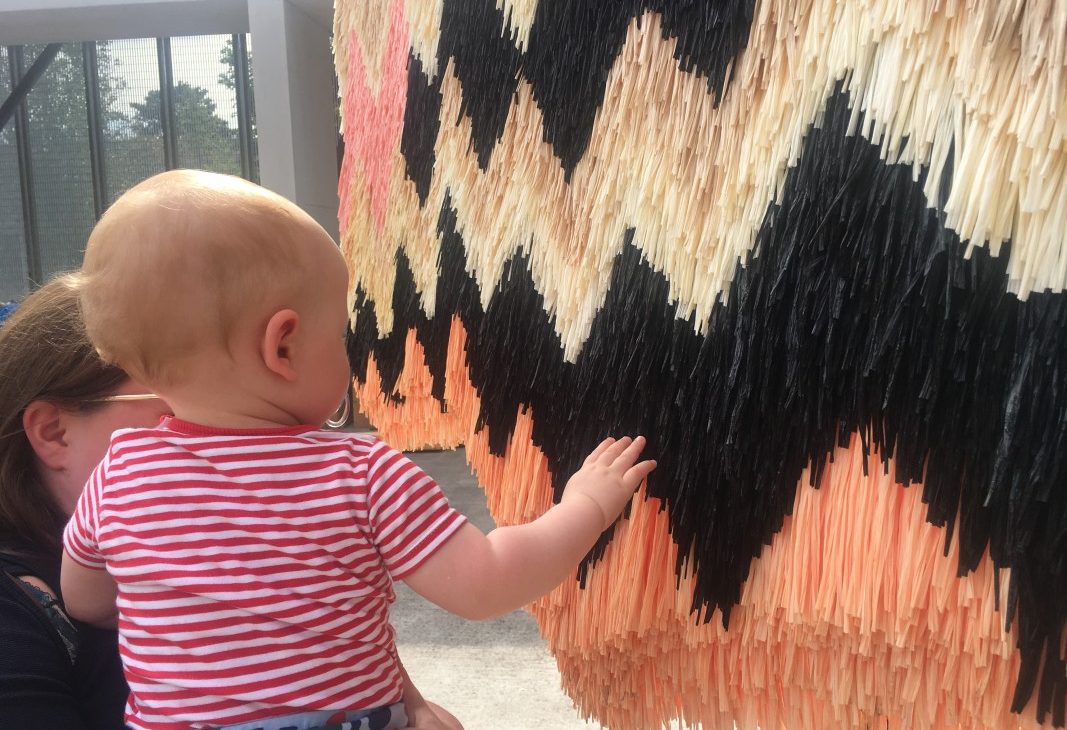 A small child in a red and white striped top is held at shoulder height, they reach toward a section of a tufted sculpture made out of pink, black, white and brown plastic