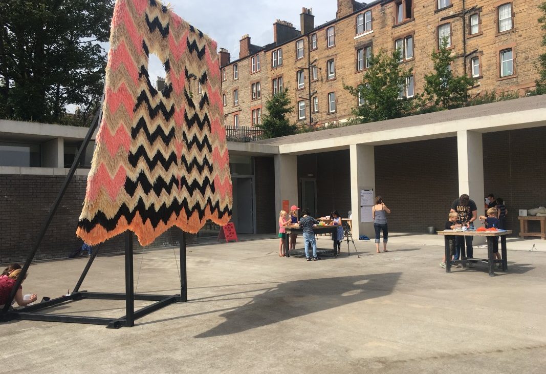 An owl like tufted sculpture with two eye holes, has a zig zag pattern in pink, cream, white black and orange. It is held up on a black metal armature in an open courtyard. There are tables spaced around with children taking part in workshops