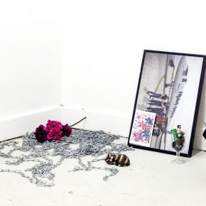 View of a corner with objects on the floor, components inclue: a silver chain, pink and purple flowers, wine glass with batteries, plastic cat, a framed picture and postcard leaning against the wall, a glass lantern with a bag draped on it