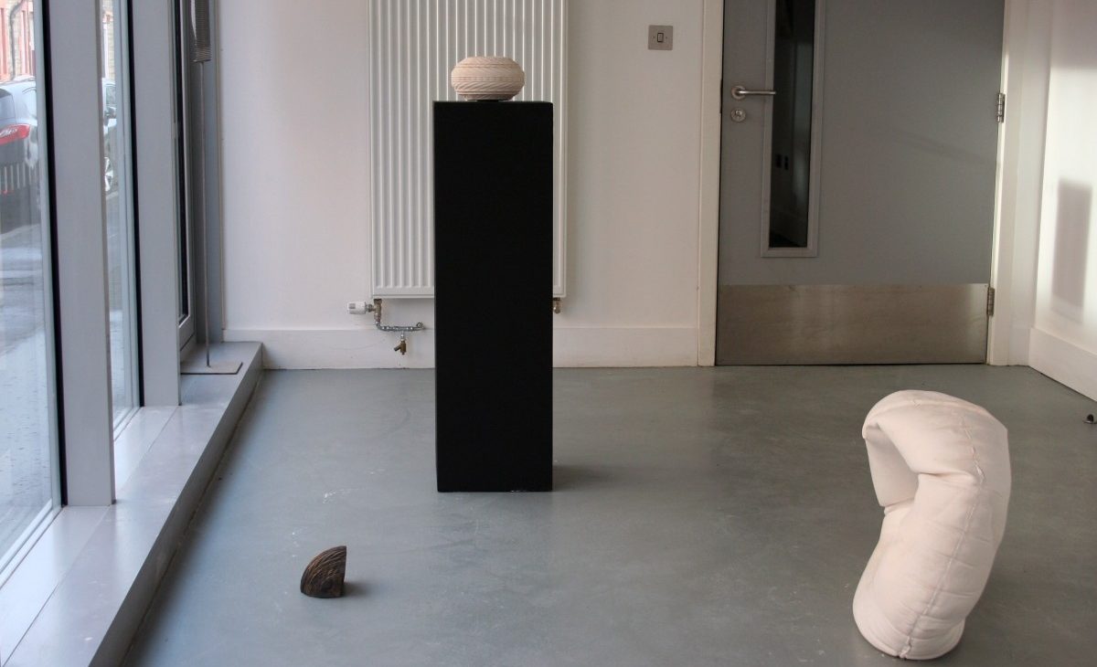 Installation view of a plaster cast rubber ring on the floor; A white jesmonite relief patterned sculpture on a black plinth with a fragment of the same sculpture cast in bronze on the floor