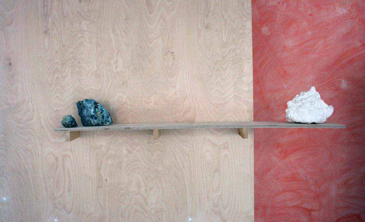 Detail of a birch plywood shelf on a freestanding plywood sheet with a plaster cast rock counterweighted by two rocks. The plaster cast is white against a washed out terracotta coloured wall