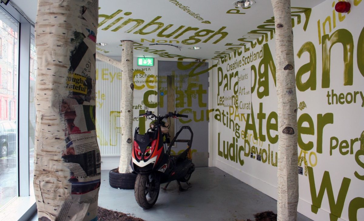 Detail view of three paper mache silver birch tree trunks sitting in dirt and bark, with a red, white and black motorcycle in the background, surrounded by alternate sizes of text painted in green on the white walls and ceiling and the grey door, excerpts include: Edinburgh, neomedievalism, Ludic, Creative Scotland, craft, creative, curating, learning