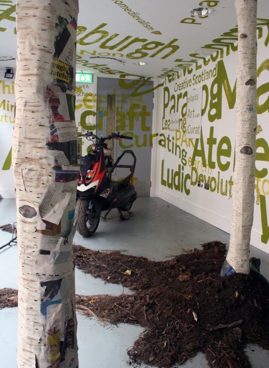Detail view of two paper mache silver birch tree trunks sitting in dirt and bark, with a red, white and black motorcycle in the background, surrounded by alternate sizes of text painted in green on the white walls and ceiling and the grey door, excerpts include: Edinburgh, neomedievalism, Ludic, Creative Scotland, craft, creative, curating, learning