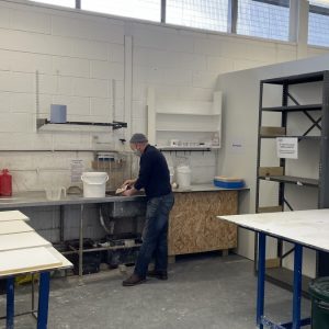 A view across the Plaster Workshop at ESW with a figure working at the far side, a worktable on the right, another work top on the left and shelving.