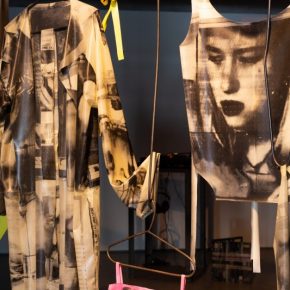 Blacksmith forged coat hangers and hooks hang from the ceiling holding white and pink latex garments, each with collaged black and white images of zoomed in faces and profiles