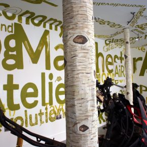 Two paper mache silver birch tree trunks are surrounded by motorcycle parts, leaves and dirt. Alternate sizes of text are painted in green on the white walls and ceiling, excerpts include: Atelier, Scotland, Economy, Devolution, Curate, Art, Craft, Resources, Learning, Peer, AiR, Glasgow, Glasgow Miracle, Manchester, futurity, medievalism
