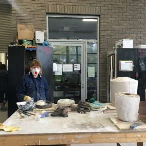 Artist Paulina Sandberg wearing P3 mask is standing on the left side of a large work table working on plaster mould jackets, there are larger moulds sitting on the other side of the table