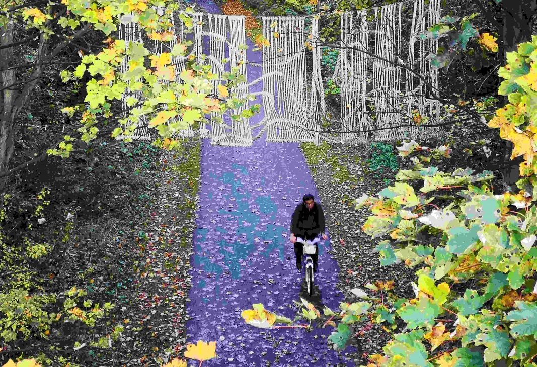 View onto cycle path with a cyclist moving underneath a macrame artwork made from ropes and knots installed at height between two trees