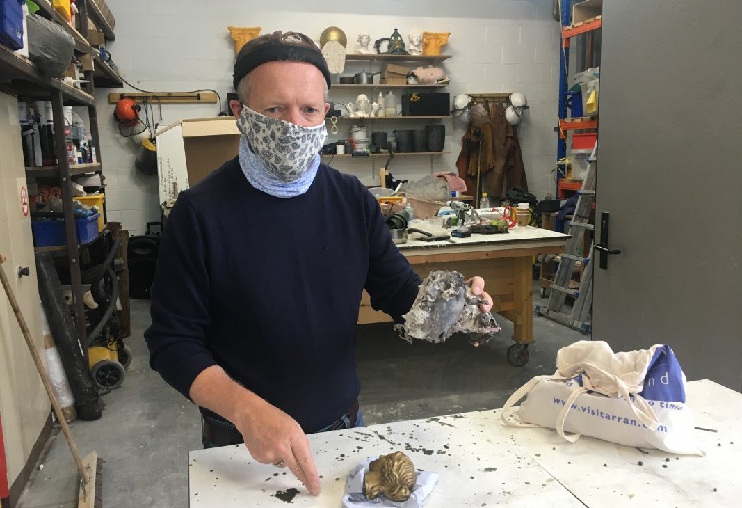 Artist Paul McAuley in a workshop holds up a bronze cast head that has remnants of sand, a smaller bronze head sits on the table in the foreground