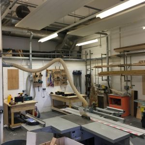 View across the Wood Workshop at ESW showing the central, large table saw and workspaces at the far side, as well as storage and other equipment along the left wall.