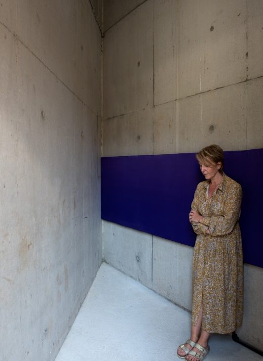Woman in dress and sandals leaning against a wall based cushioned blue ribbon listening to sound installation