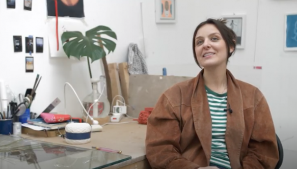 Emma Hislop sitting at her desk in her studio at ESW wearing a brown jacket and striped tshirt.