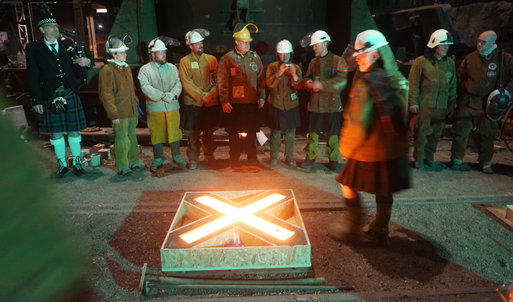 Line of people including a piper in highland dress standing alongside an X mould with glowing red metal in it.