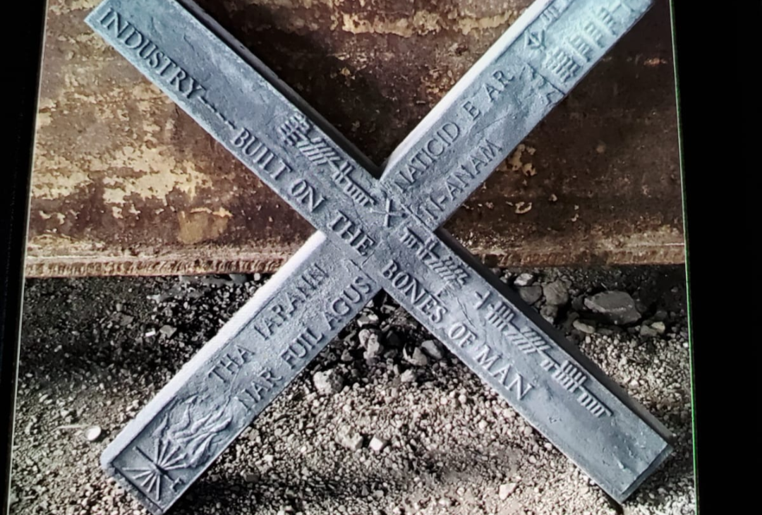 A cross sculpture, in cast metal, with phrases in English and Gaelic including 'Industry... built on the bones of man'.