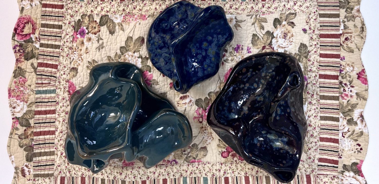 Three ceramic sculptures in deep green, blue and darker glaze on a textile mat, viewed from above.