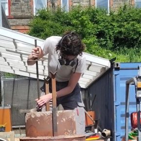 Meabh Breathnach stands above a rusty metal iron furnace preparing it in a sunny yard.