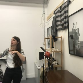 Oana Stanciu in studio during studio visit, gesturing, with objects and prints on the wall.