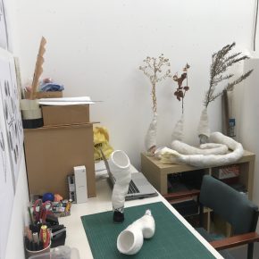 View of a desk and wall space in Andrew Gannon's studio with a green cutting mat and several plaster limb sculptures alongside dried foliage.