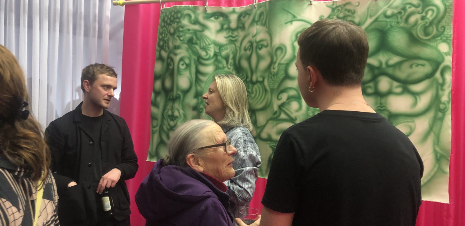 Four people talking on front of Suds McKenna's green, figurative, spray painted work on canvas, hanging on front of the pink curtain within Dissenter Space's set up.