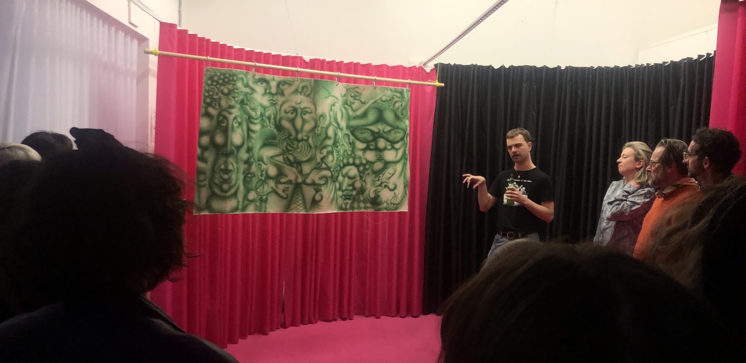 Suds McKenna standing to the right of their hanging spray painted work, within the black and pink curtains of the Dissenter Space set up.