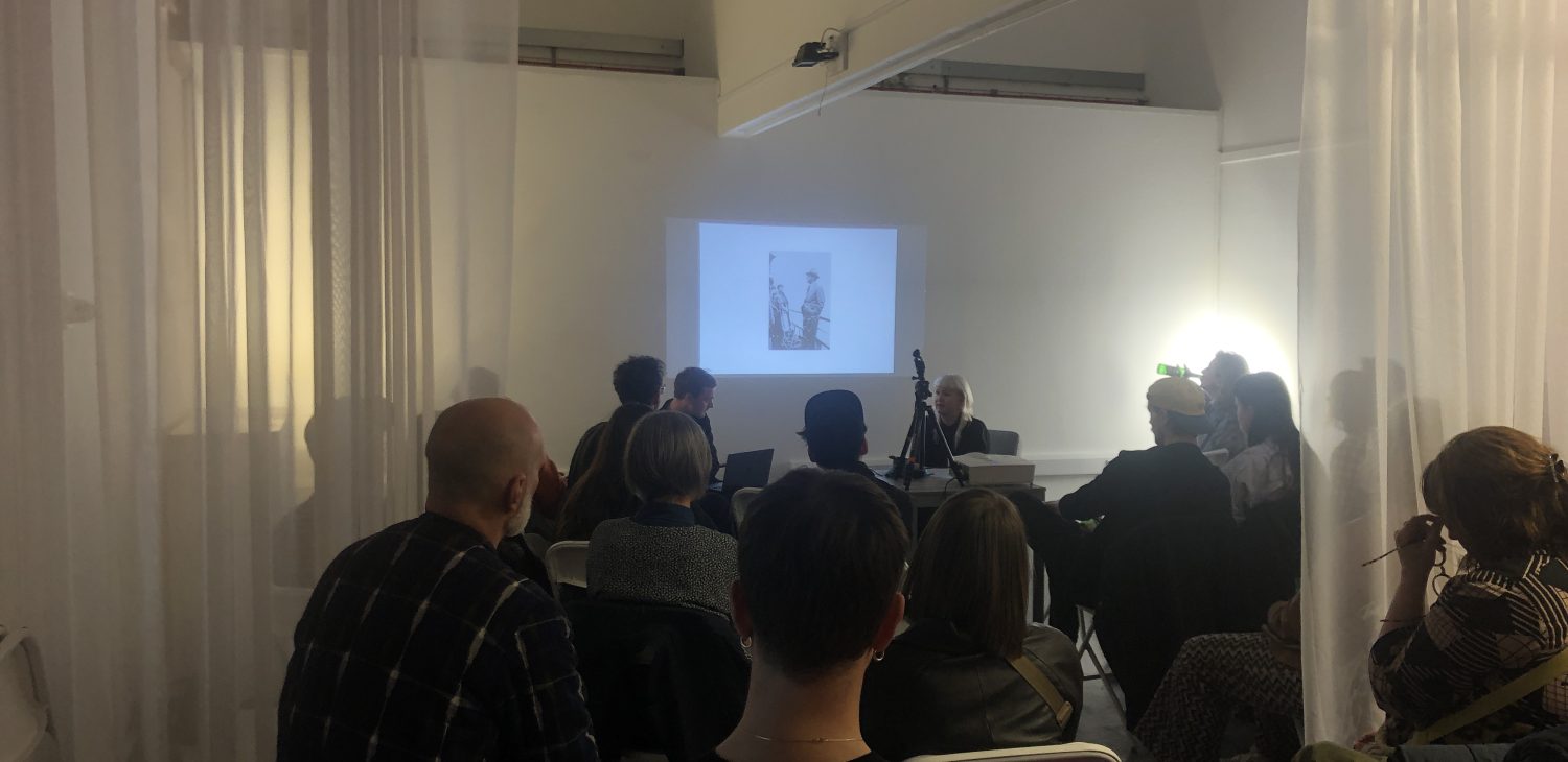 Duo WoodcockEllis presenting to the audience of a Dissenter Space talk within a white room with translucent white curtains.