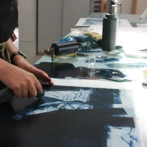 Predominantly blue prints on paper are laid out on a desk top with the participant's hands visible to the left.
