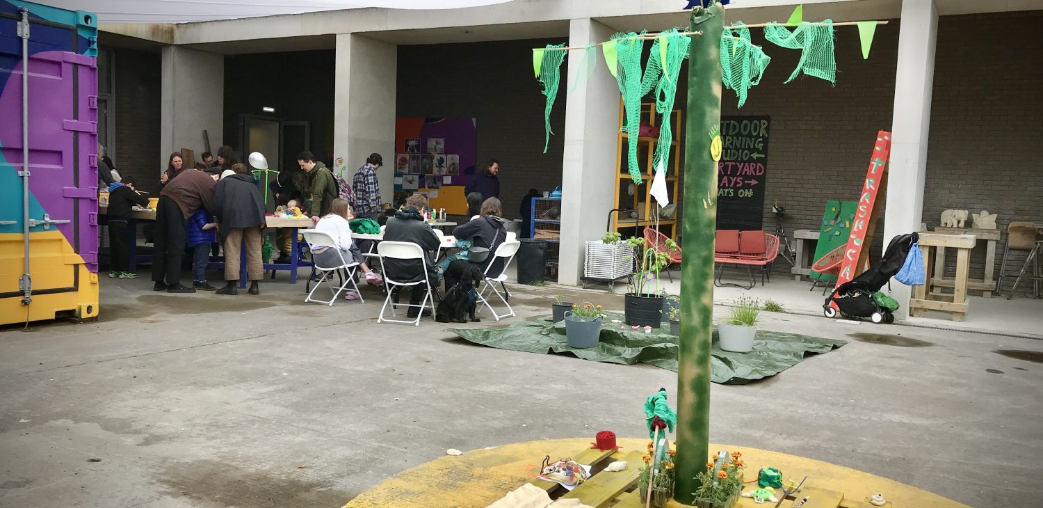 A desert island and palm tree made from old materials in the foreground and people working as part of a Sculpture Saturdays workshop in the background.