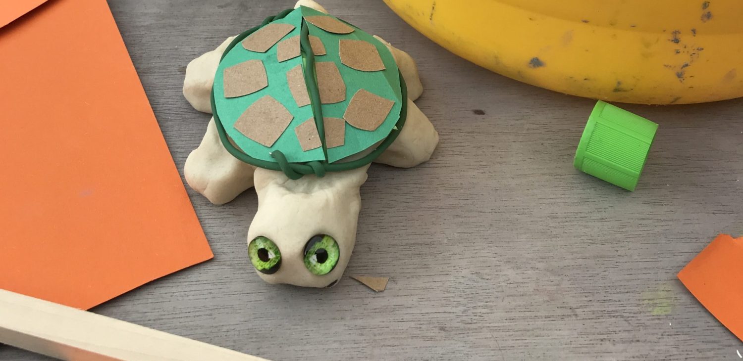 A small green and beige tortoise sculpture on a work table with orange paper and a yellow canister next to it.