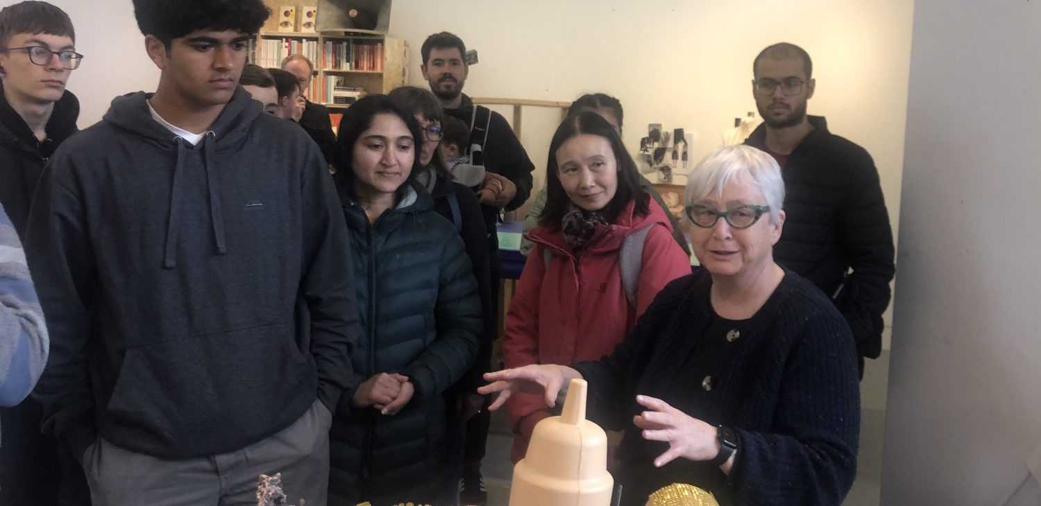 Tour visitors speak to Project Space Holder Jan Pimblett, standing around her work table which has objects she is making and integrating into assemblage sculptures.