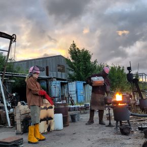 Outside view with two people tending a flaming furnace wearing protective equipment. One figure is pouring oil into the mechanism.