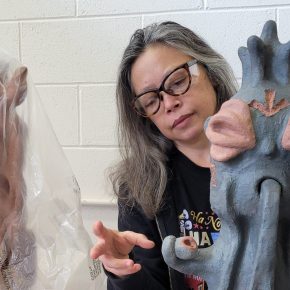 Mai Nguyễn-Long working on a dark clay sculpture with another covered in plastic sheet alongside.