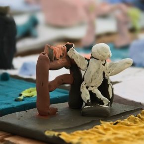Close-up of a handmade clay sculpture by P5 pupils from Victoria Primary School depicting two human-like figures in action, one white figure seated or leaning against a dark structure while a reddish-brown figure arches over it, set on a textured base.